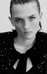 Close-up portrait of a model with dark hair wearing a black sequined top against a textured background.