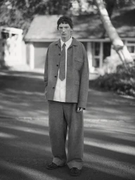 Monochrome portrait of a young male model standing on an asphalt street wearing a loose grey jacket, matching trousers, tie, and white shirt.