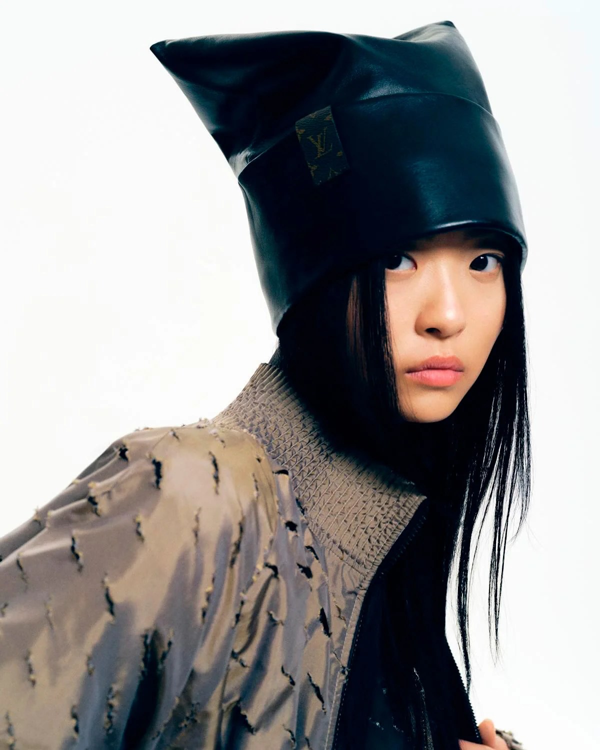 Fashion shot: Young Asian woman wears a black leather hat with logo, taupe jacket, posing against a white backdrop.
