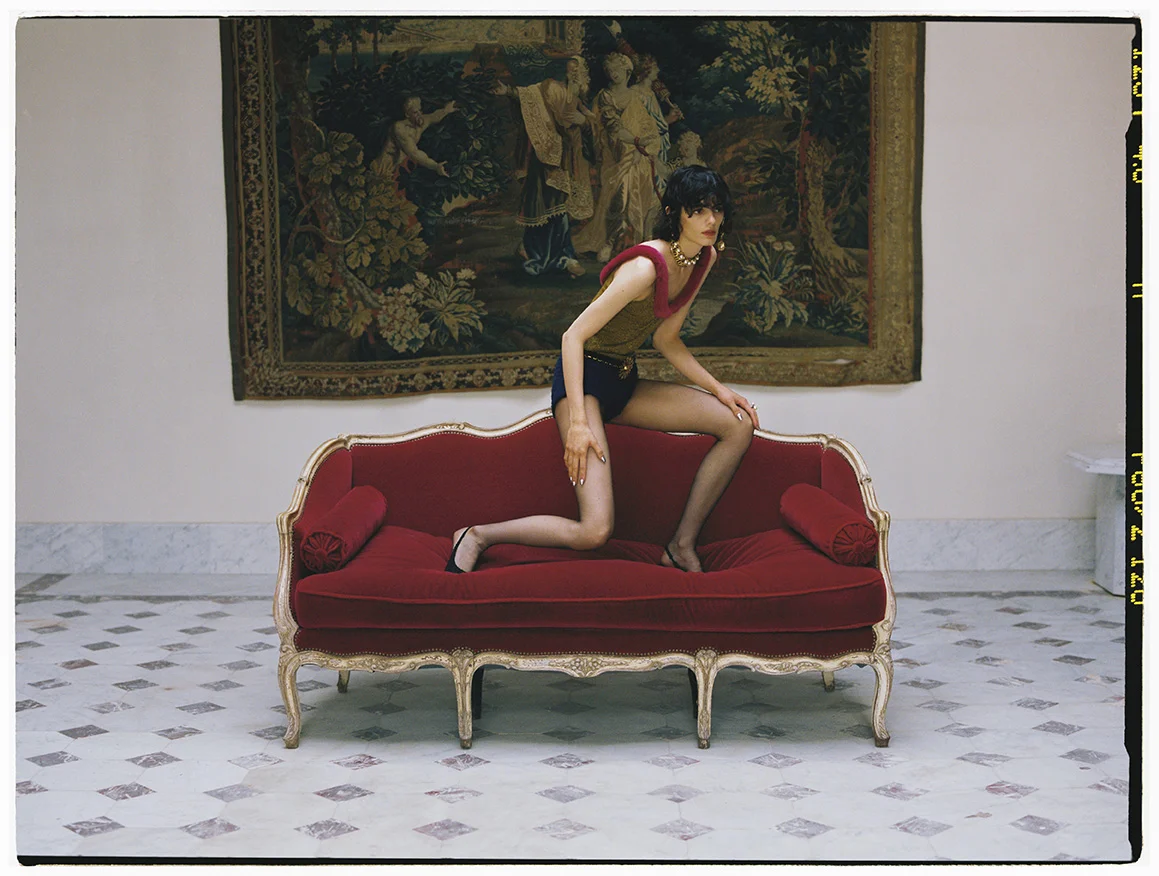 A fashionable woman poses on a red couch in an elegant indoor setting with a tapestry, wearing a tan top and black shorts.