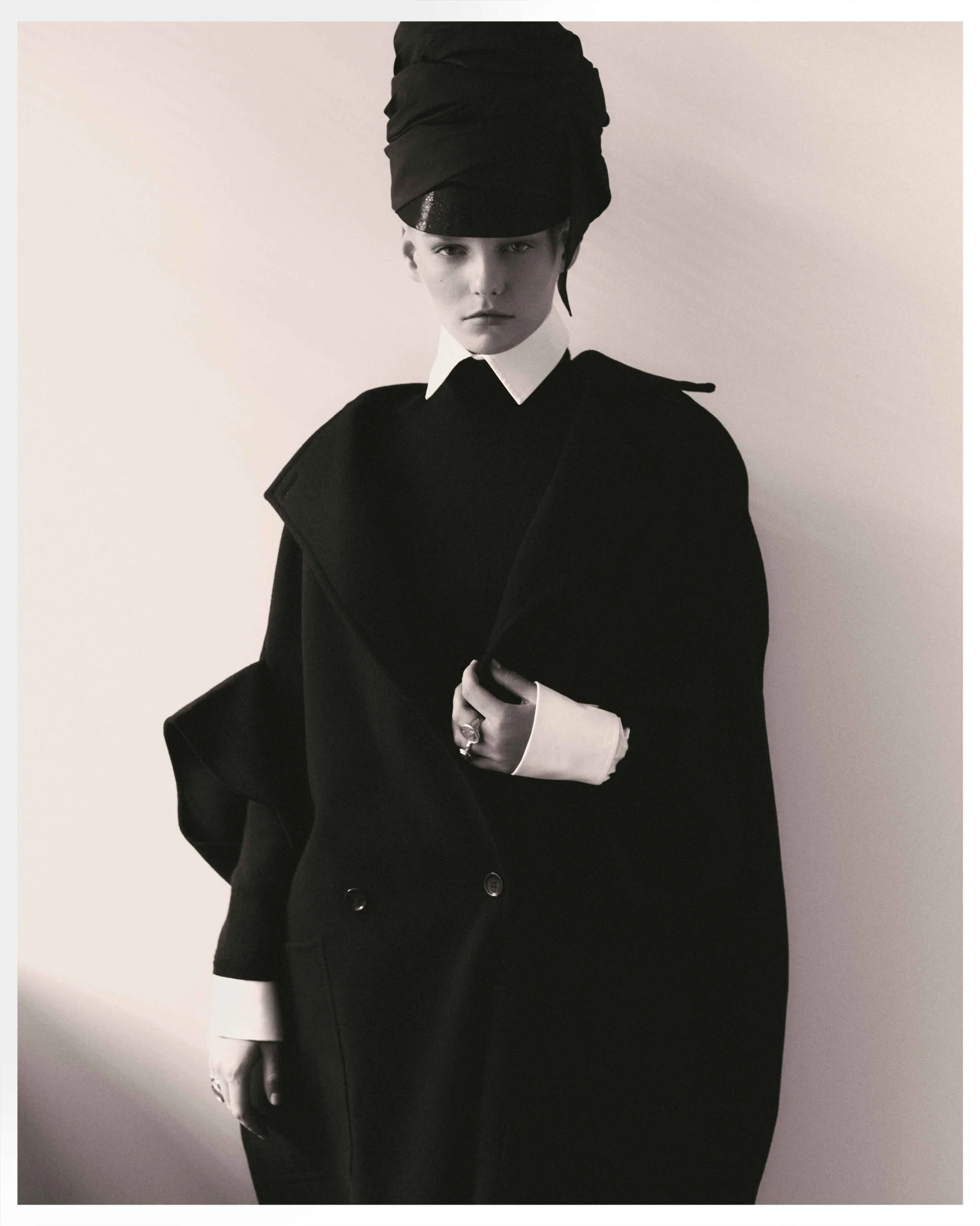 Monochrome portrait of a young woman in a black overcoat and matching head wrap against a light neutral background, fashion photography.