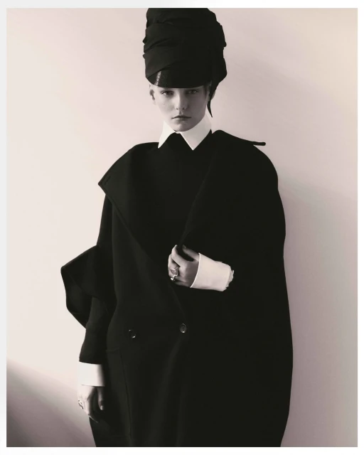 Monochrome portrait of a young woman in a black overcoat and matching head wrap against a light neutral background, fashion photography.