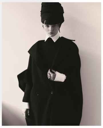Monochrome portrait of a young woman in a black overcoat and matching head wrap against a light neutral background, fashion photography.