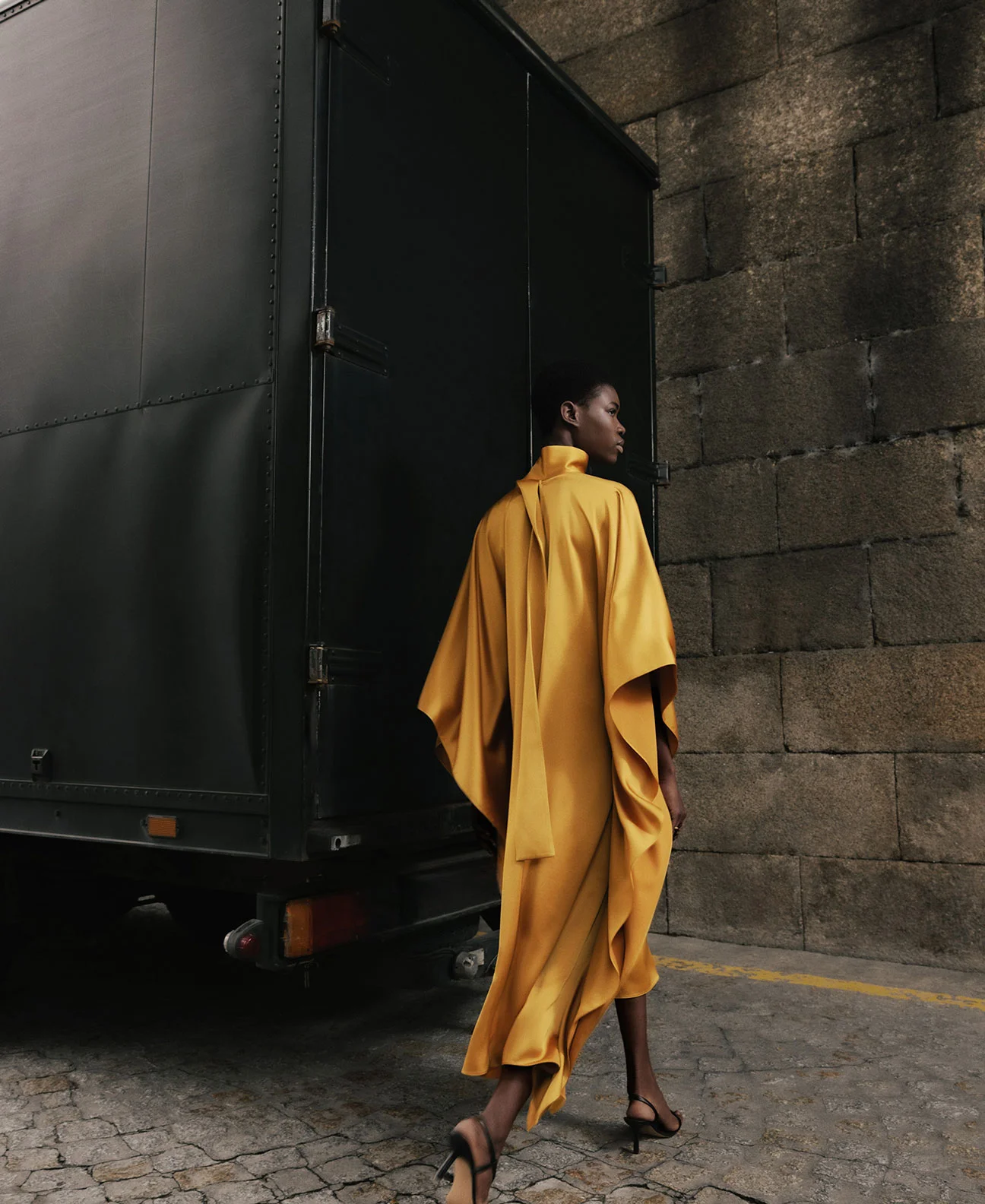 An African-American female model wearing a yellow dress walks past a dark trailer and stone wall in an outdoor fashion shot.
