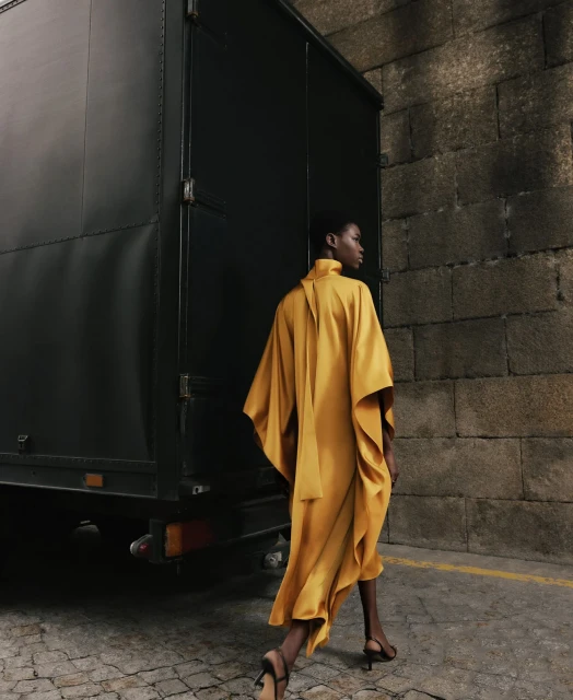 An African-American female model wearing a yellow dress walks past a dark trailer and stone wall in an outdoor fashion shot.