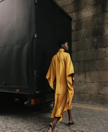 An African-American female model wearing a yellow dress walks past a dark trailer and stone wall in an outdoor fashion shot.