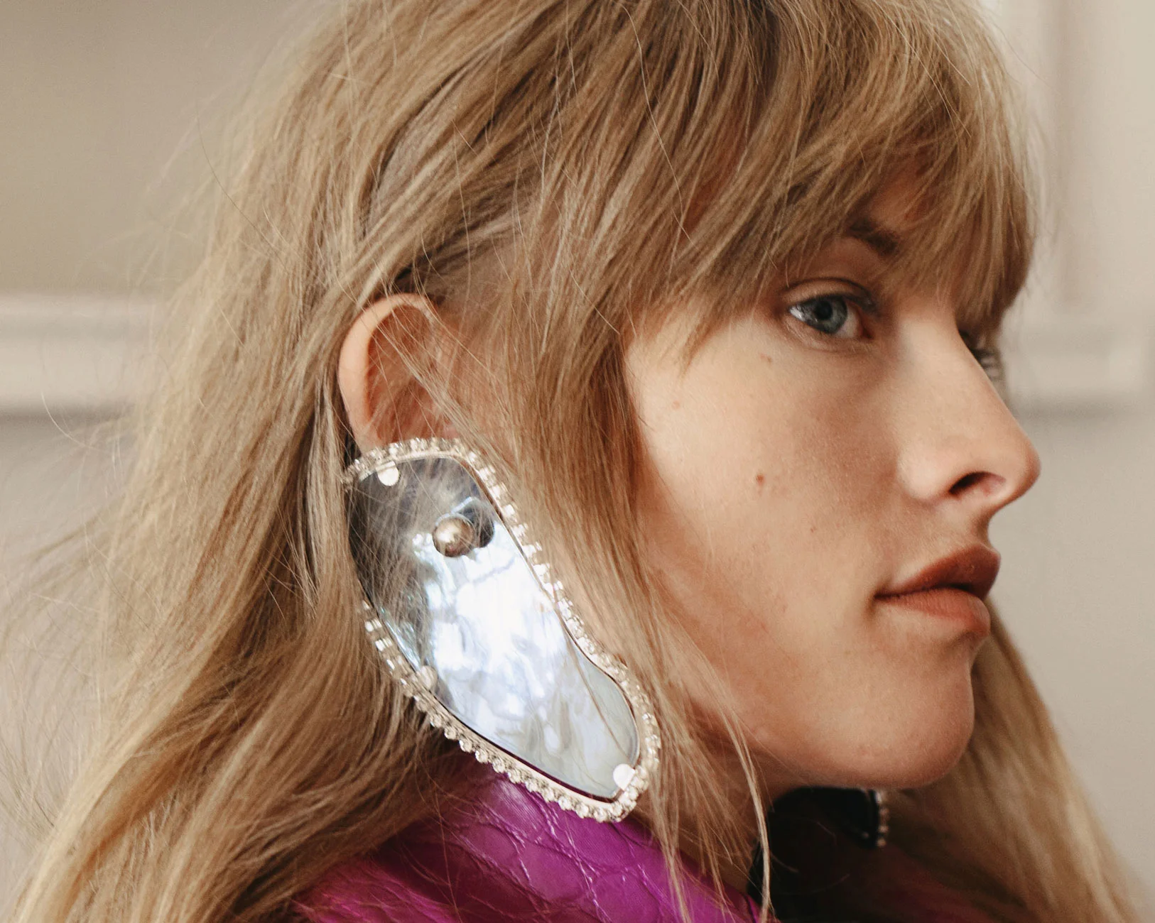 Blonde woman's portrait wearing a purple snakeskin jacket and a large rhinestone and silver statement earring in a studio setting.