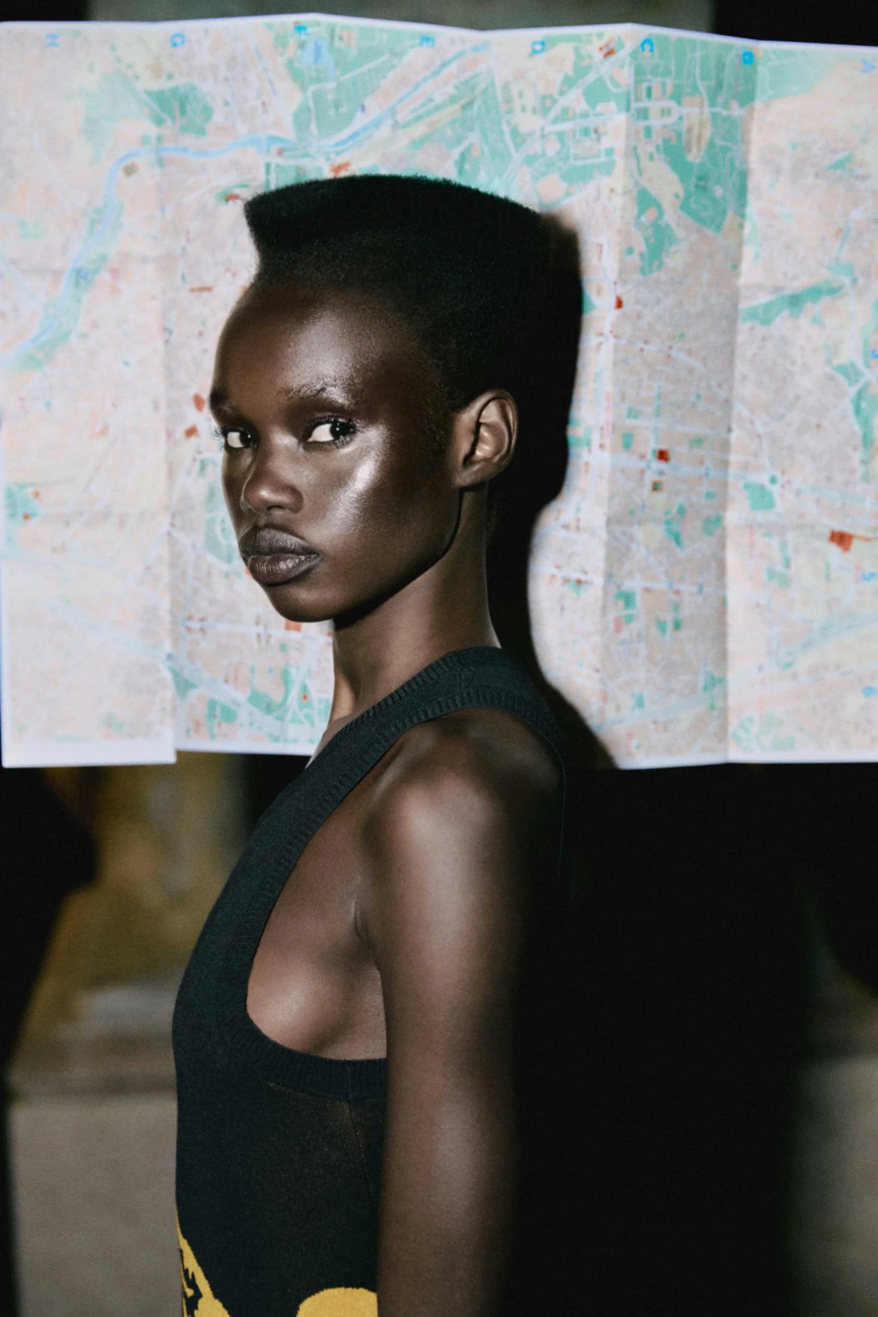 Profile of an adult woman with dark skin and a high-top fade, wearing a black tank top and posing in front of a map backdrop.