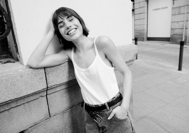 Smiling woman in white tank top and denim jeans leaning against a stone building outdoors