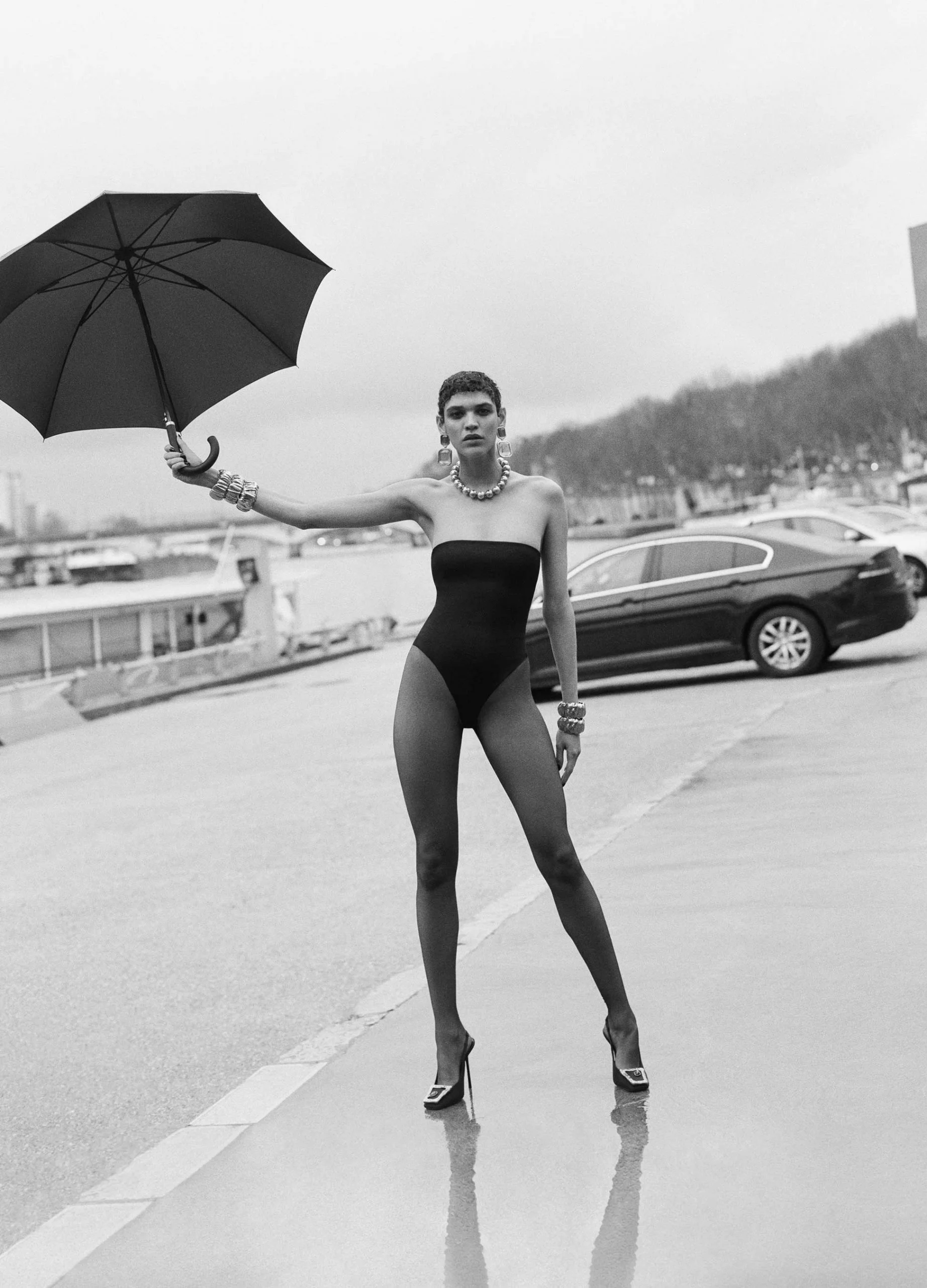 Black and white fashion shot of a woman in a black bodysuit holding an umbrella, with city buildings and a car in the background.