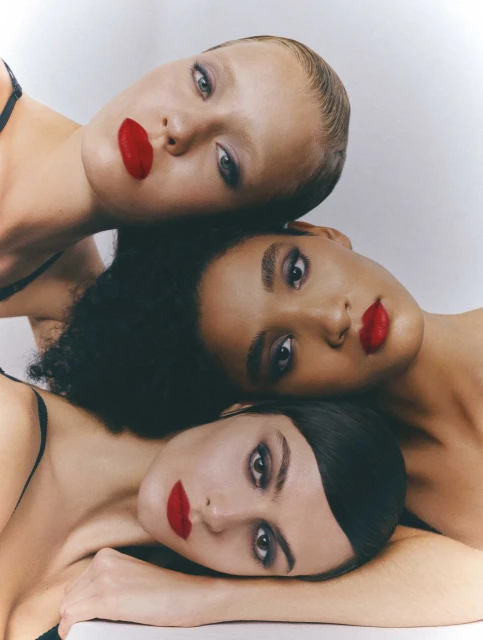 Close-up studio portrait of three diverse young adult women with red lipstick, dark eye makeup, fair skin, and black tops.