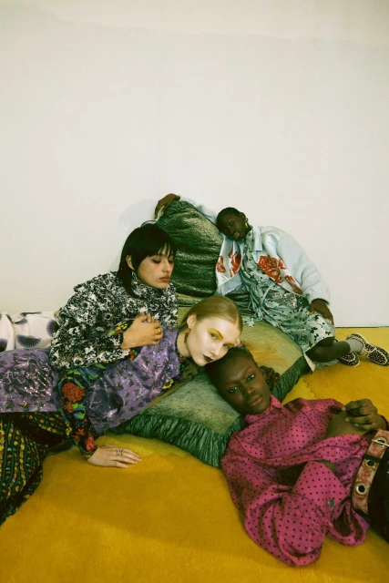 Four models pose in colorful patterned outfits against a white wall, lying on yellow and green cushions, fashion editorial.