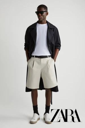 A man wearing dark sunglasses, a black coat over a white t-shirt, light trousers, and white sneakers against a plain background.