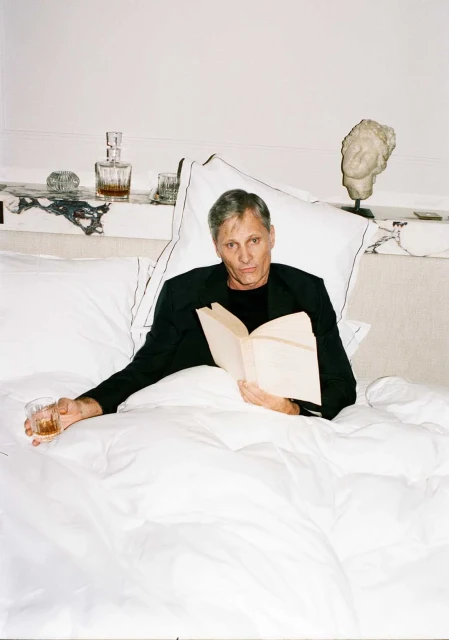 Viggo Mortensen relaxes in bed with a book and a drink, dressed in a black blazer, in a bright, indoor setting with a white interior.