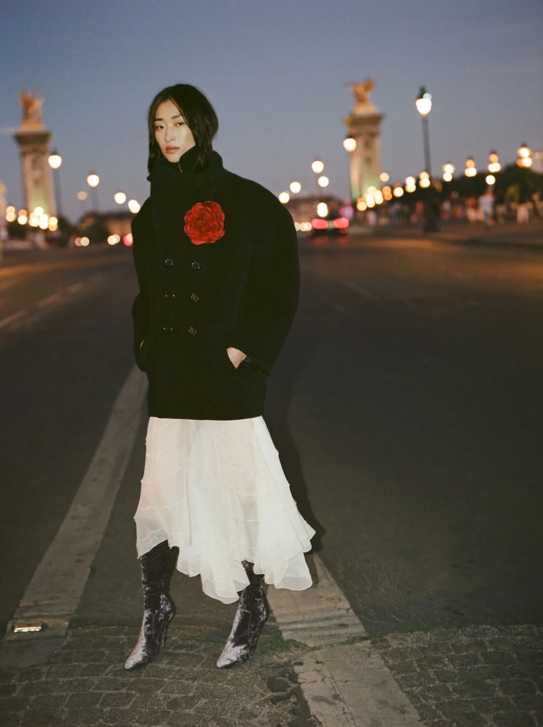 Elegant woman in a black coat with a red flower detail and a white skirt poses on a bridge with illuminated architecture at night.