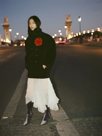 Woman in dark coat with a red flower pin, white skirt, and boots standing on a city street at dusk.
