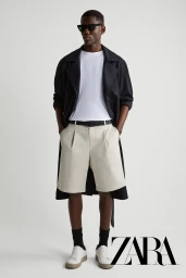 A man wearing dark sunglasses, a black coat over a white t-shirt, light trousers, and white sneakers against a plain background.