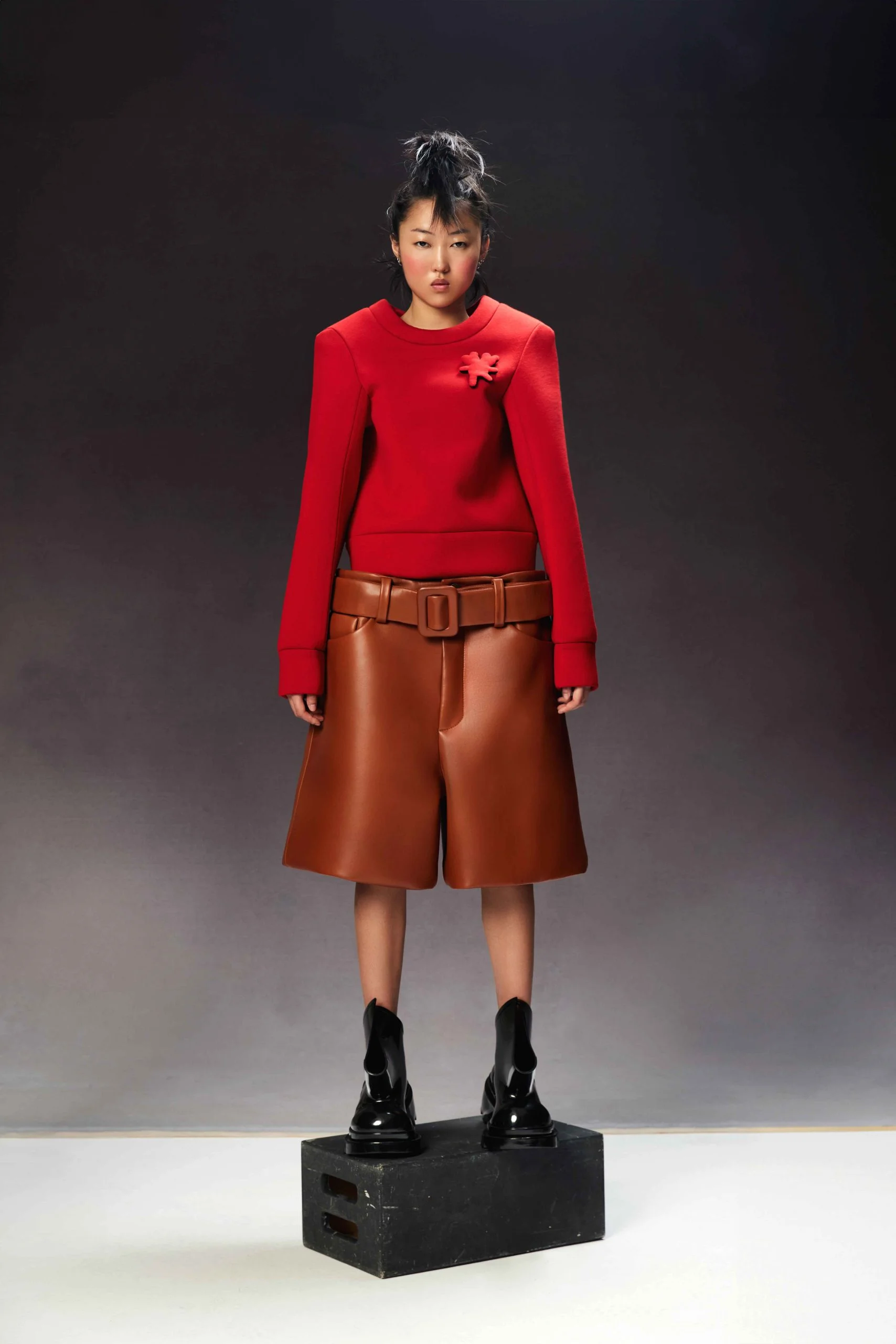 A full-length portrait of a young Asian woman in a red top and brown shorts standing on a black cube against a gray backdrop.