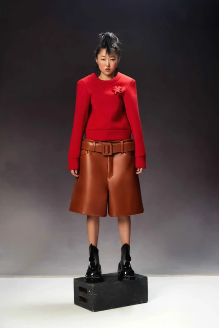 A full-length portrait of a young Asian woman in a red top and brown shorts standing on a black cube against a gray backdrop.