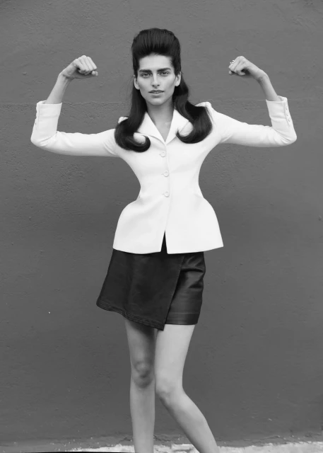 Black and white studio shot of a slender woman with retro hair flexing her biceps while wearing a white jacket and black skirt.