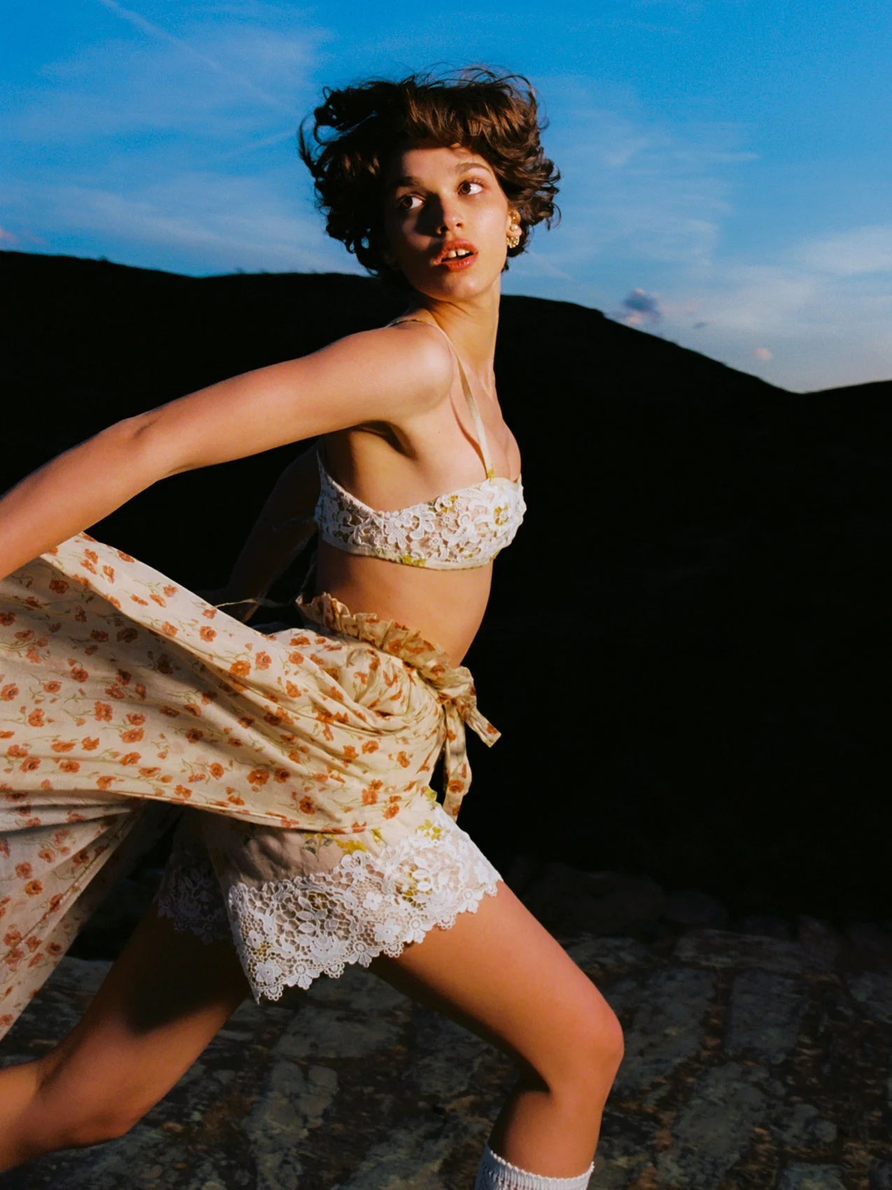 Young woman in a floral bralette and skirt runs outdoors; dark hill and twilight blue sky visible in background.