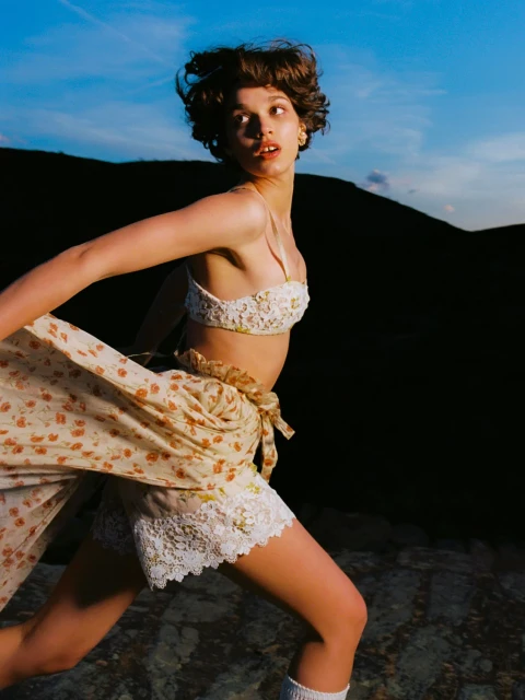 Young woman in a floral bralette and skirt runs outdoors; dark hill and twilight blue sky visible in background.