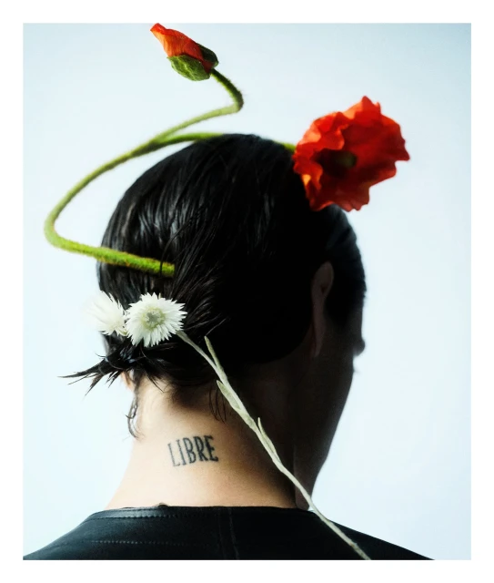 Woman with slicked-back dark hair adorned with red and white flowers, viewed from behind, featuring a neck tattoo and a light background.