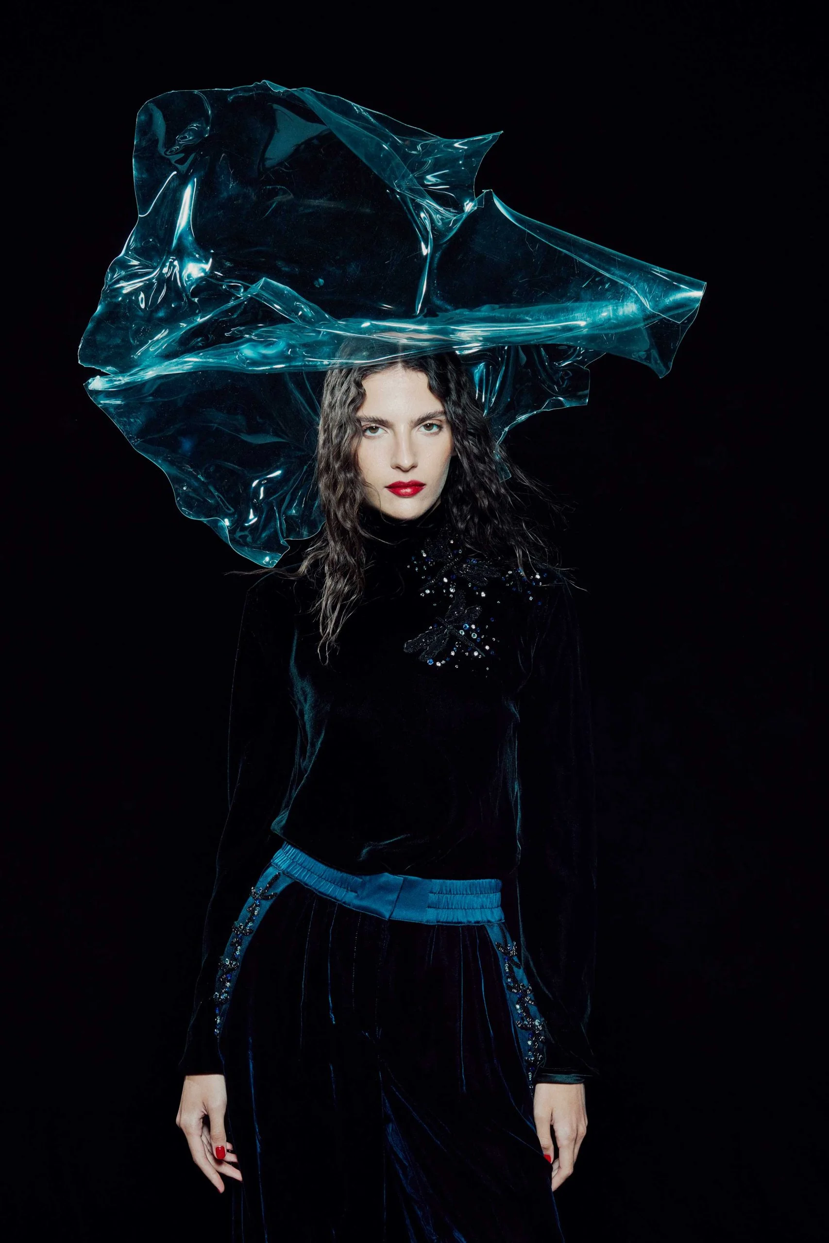 Woman wearing a black velvet top and pants with a blue transparent plastic headpiece posing in front of a black background in a studio shot.