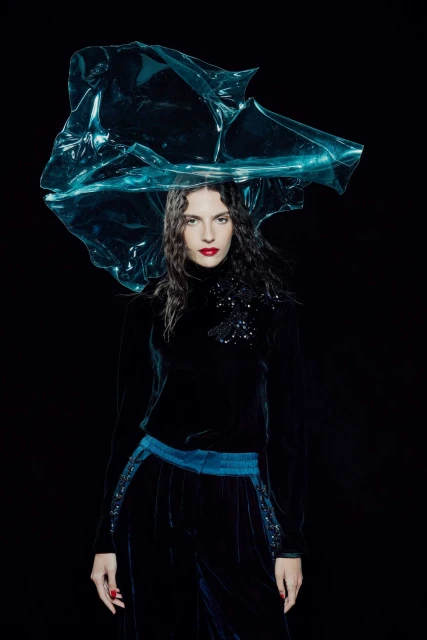 Woman wearing a black velvet top and pants with a blue transparent plastic headpiece posing in front of a black background in a studio shot.