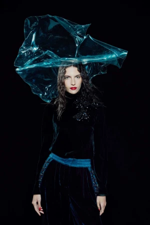 Woman wearing a black velvet top and pants with a blue transparent plastic headpiece posing in front of a black background in a studio shot.