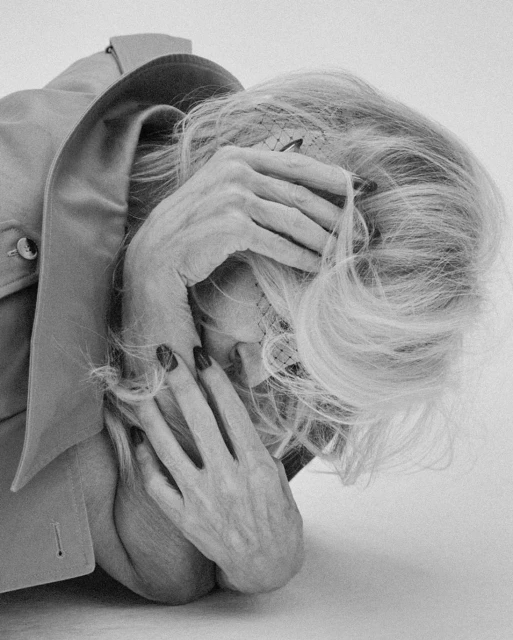 Monochrome portrait of an elderly woman resting her face in her hands with painted nails, wearing a jacket in a light studio setting.
