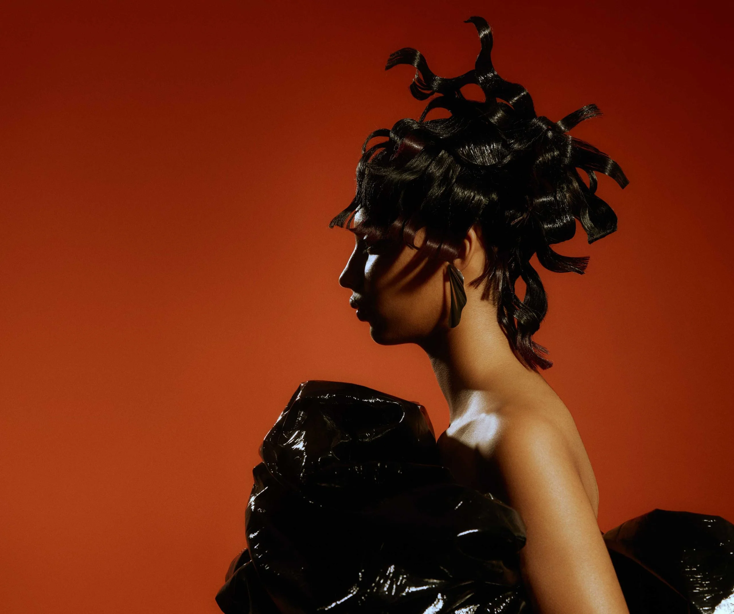Profile shot of a model with styled black hair and glossy black garment against a reddish-orange backdrop in a studio setting.