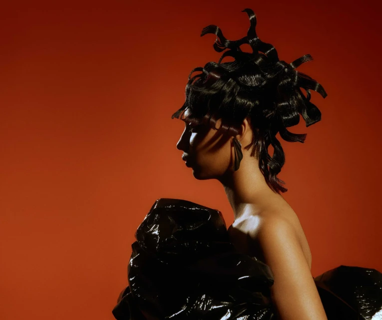 Profile shot of a model with styled black hair and glossy black garment against a reddish-orange backdrop in a studio setting.