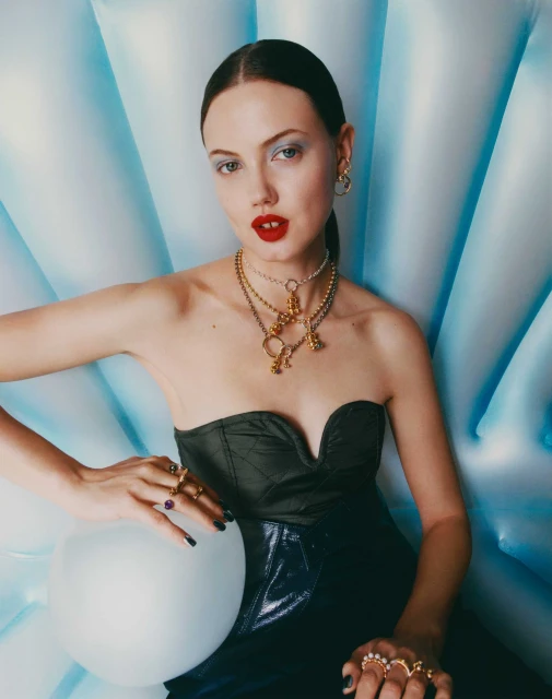 Close-up fashion shot of a young adult woman wearing an olive bustier, dark skirt, jewelry, and make-up, posing in front of a blue shell.