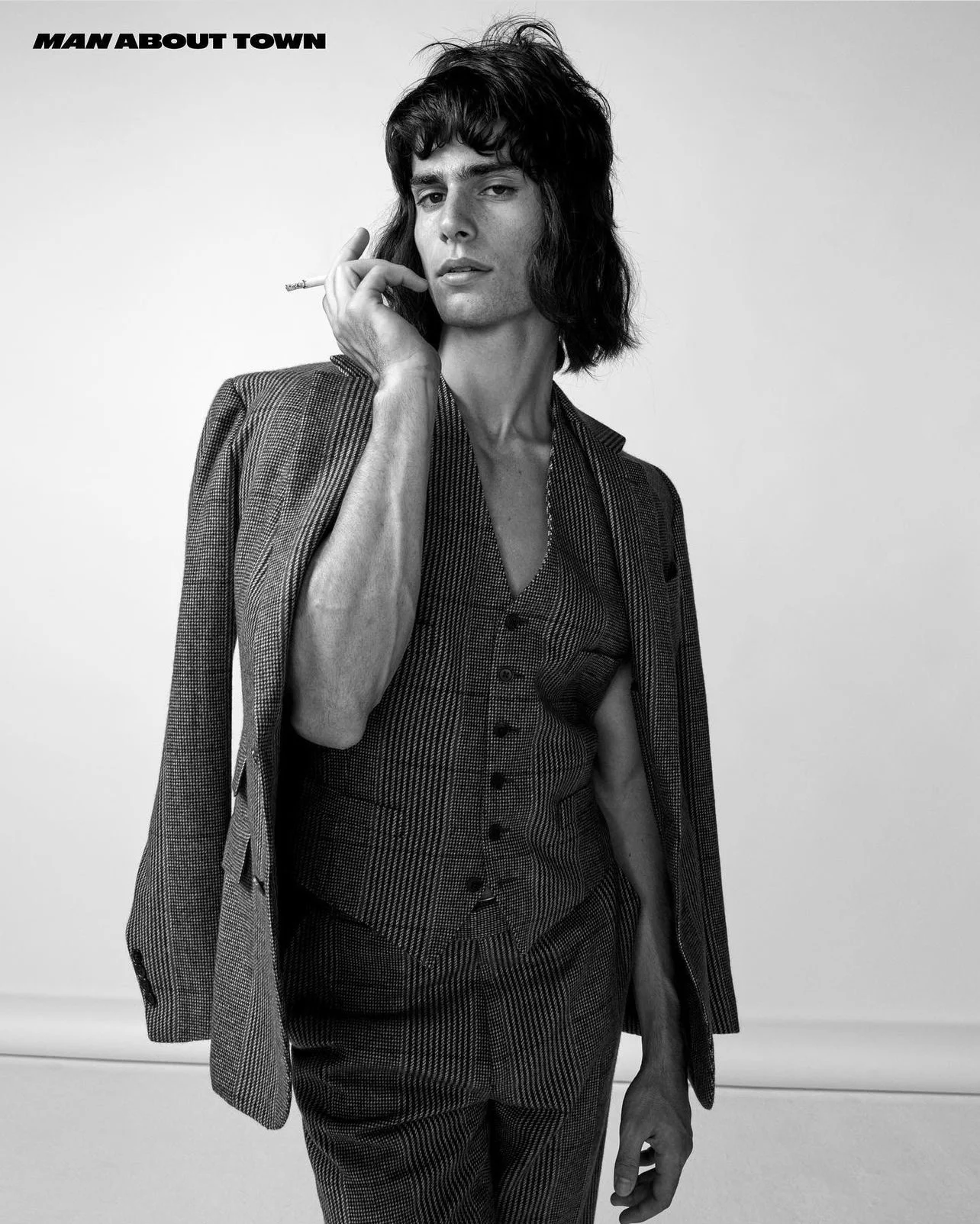 A man in a striped suit poses in a studio, holding a cigarette to his mouth, against a neutral backdrop, black and white.