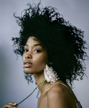 Portrait of a black woman with voluminous curly dark hair holding a pale rose flower.