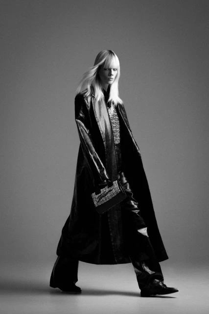 Black and white fashion shot of a slender adult female posing in a long black coat and black pants, holding a black clutch.