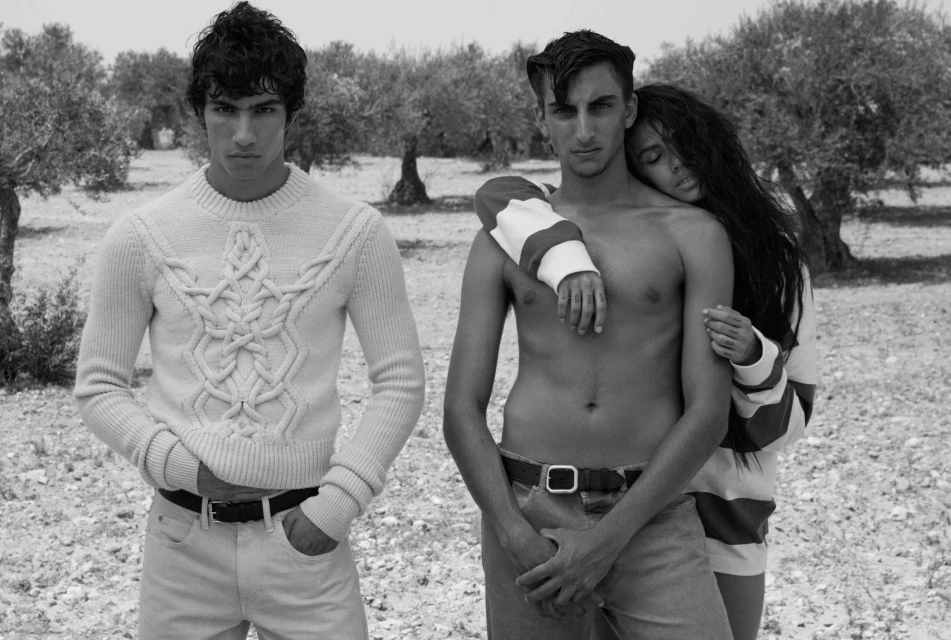 Black and white shot of three people outdoors, one wearing a sweater, embracing in a field near trees. Fashion or editorial style photo.