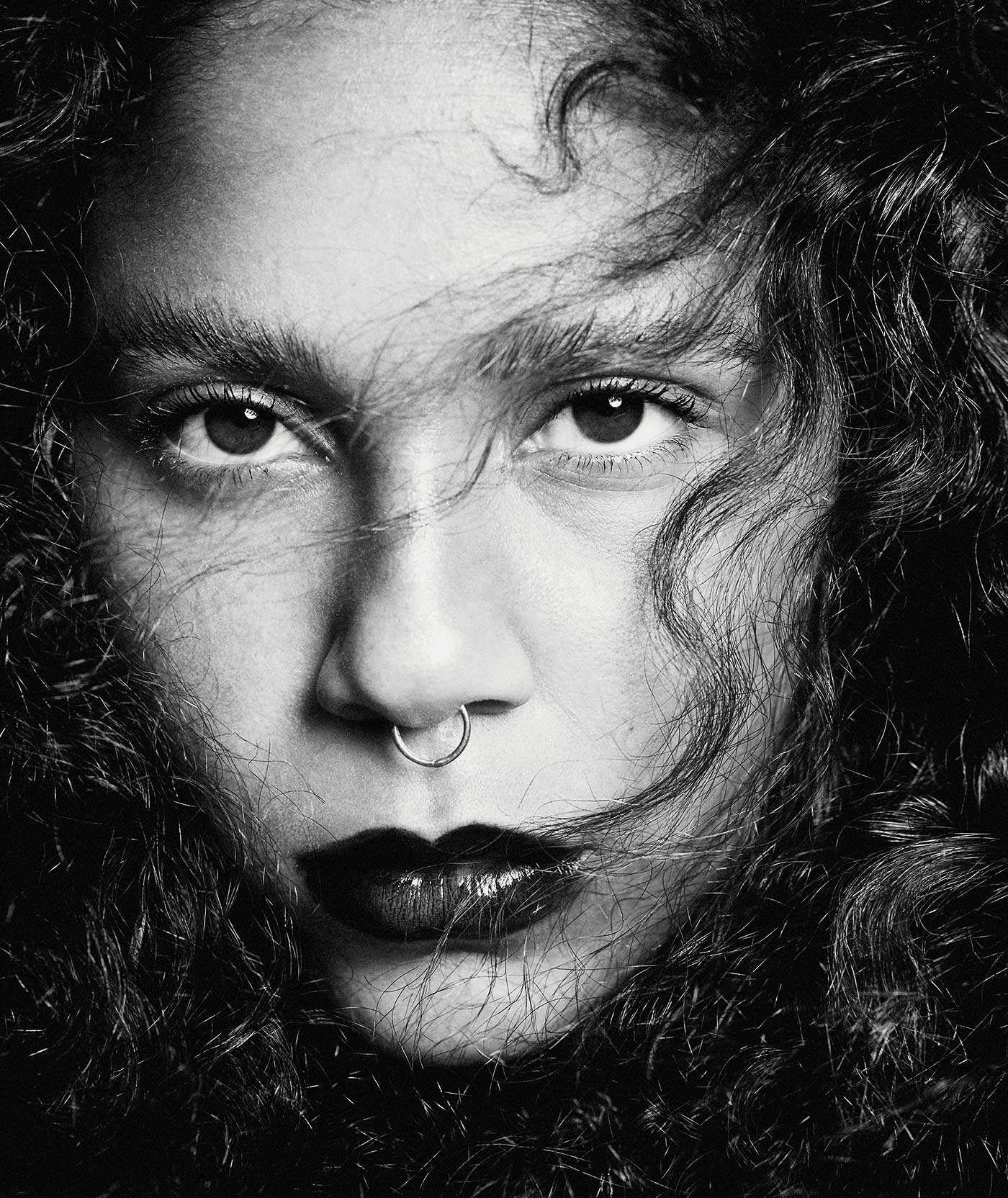 Monochrome portrait of a young woman with dark curly hair, a nose ring, and dark lipstick staring intently at the camera.
