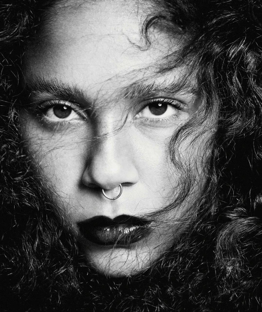 Monochrome portrait of a young woman with dark curly hair, a nose ring, and dark lipstick staring intently at the camera.