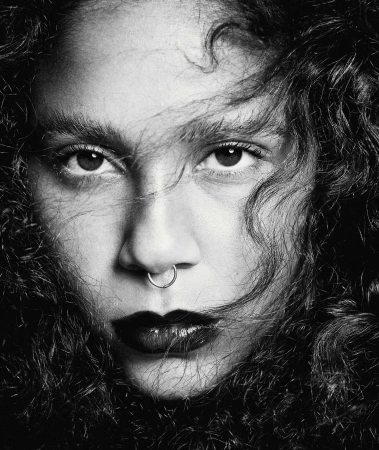 Monochrome portrait of a young woman with dark curly hair, a nose ring, and dark lipstick staring intently at the camera.