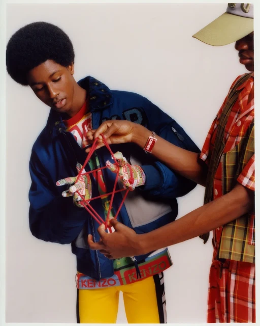 Two dark-skinned young adult models play cat's cradle with red string in a studio setting, wearing Kenzo branded apparel.