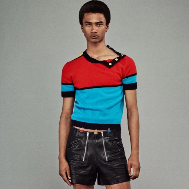 Portrait of a black male fashion model in a colour-blocked red and blue sweater and black leather shorts, standing in a photo studio.