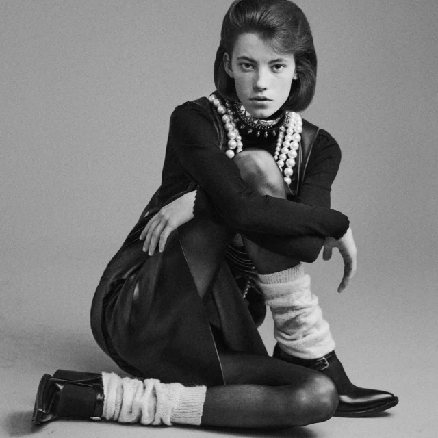 Monochrome portrait of a woman with short brown hair wearing a turtleneck, vest, pearls, and leg warmers, sitting in a photo studio.