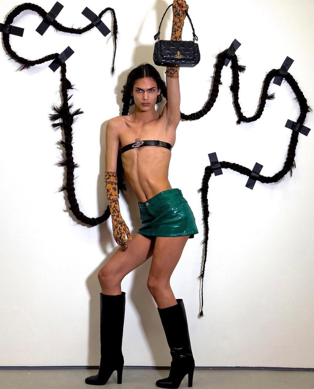 Fashionable person poses in a glossy green skirt, black boots, and harness while holding a purse and wearing arm coverings.