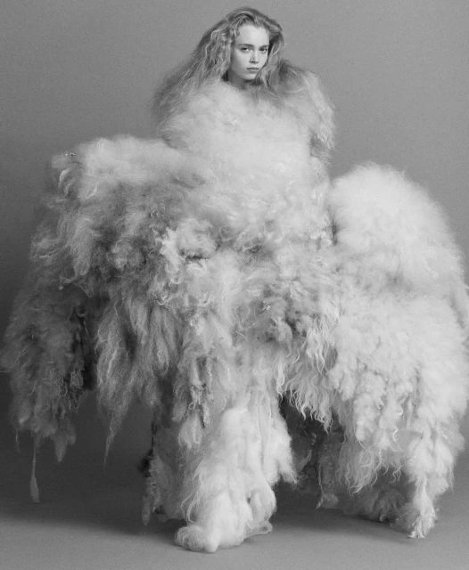 Monochrome portrait of a young woman wearing a voluminous dress made of fluffy white material, set against a plain gray background.