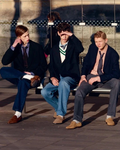 Three stylish young men are sitting on a bench in the sunlight, wearing blazers, dress shirts, ties, and loafers, looking fashionable and cool.