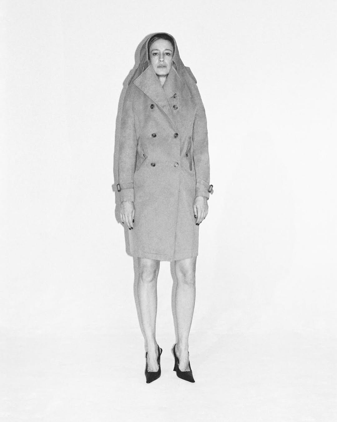 Woman in gray coat and black heels stands on white background in a studio setting, full shot, fashion editorial image.