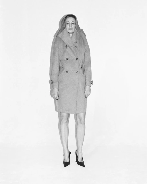 Woman in gray coat and black heels stands on white background in a studio setting, full shot, fashion editorial image.