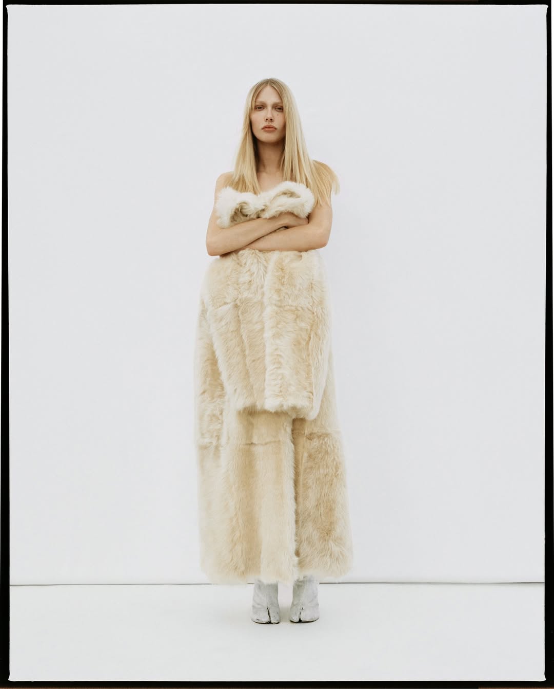Fashion shot of a blonde woman posing indoors in a large beige fur coat, wearing beige boots with a white backdrop.